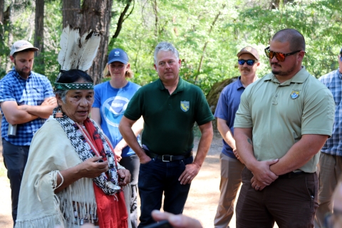 A woman in traditional Native American clothing speaks with several people wearing federal- or state-agency shirts and hats