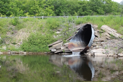 a metal culvert emerging from the side of a bank with water pouring from it into stream