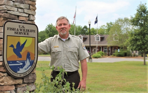 A man in a U.S. Fish and Wildlife Service uniform standing next to a pillar with a U.S. Fish and Wildlife Service logo and in front a visitor center building