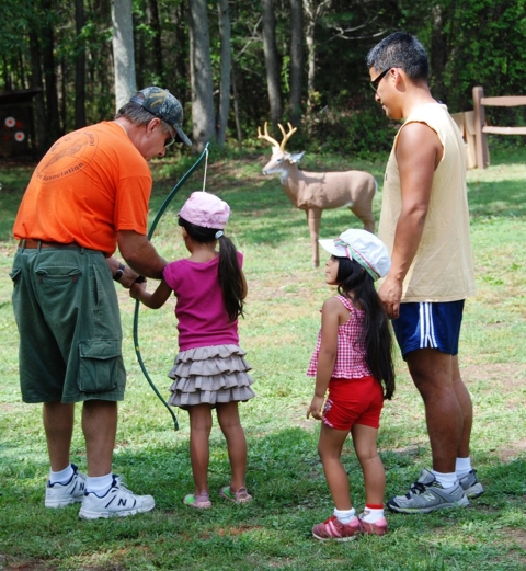 Man helping young girl learn to hold a bow. A man and daughter are watching. In the distance a manakin of a deer is set up.