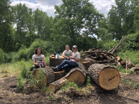 Staff members eat lunch while sitting on large logs.