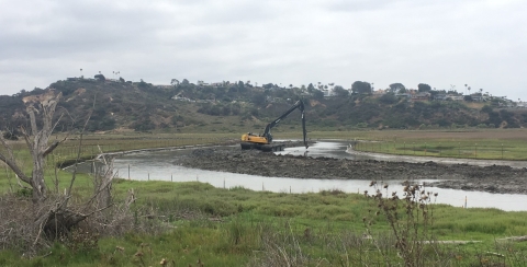 yellow excavator sits on top of sediment in lagoon