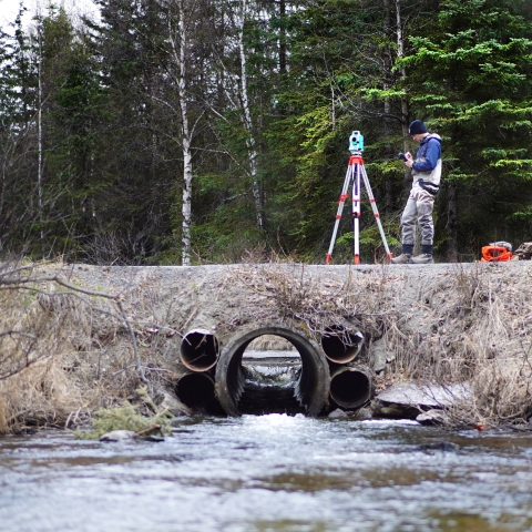 Man writing notes on a small notebook while using survey equipment on a road with an outdated culvert system under it.