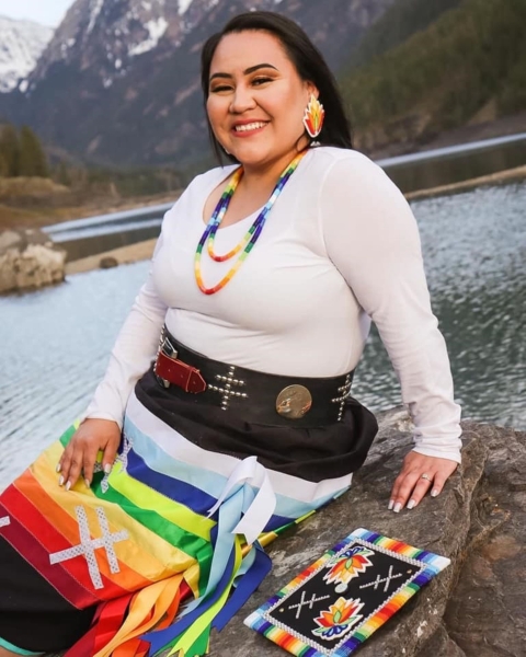  A Native American woman sits on a rock in cultural regalia with the mountains and a lake in the background