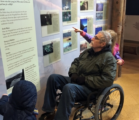 Grey-haired man in wheelchair reads interpretive sign while a girl points to it.