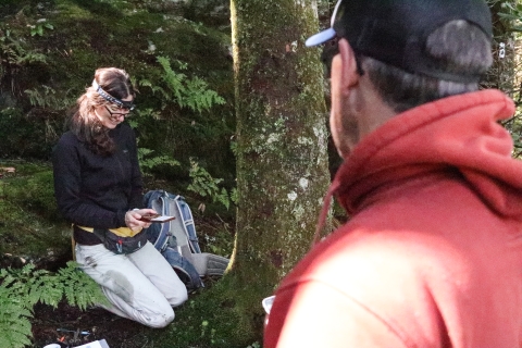 Two people in a forest. One is sitting on their knees, looking at a device in their knees. The second person is looking at the first.