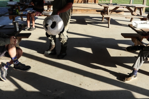 Four picnic tables, three people sitting a picnic tables, one person standing, and holding a panda bear mask