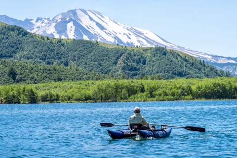 Person in small raft floats in blue lake with forest and mountain in distance