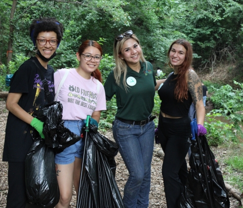 four people pose while holding black trash bags and smiling. They appear to be in a dense forest area.