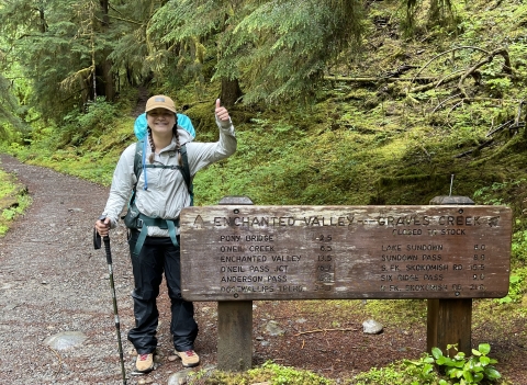 Service intern, Hannah Ferwerda, standing beside a trailhead sign in forested trail. 