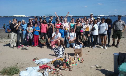 a large group of people stand smiling and waving towards the camera on the beach. a collection of items, bas and boxes sit before them on the ground. The ocean can be seen behind them