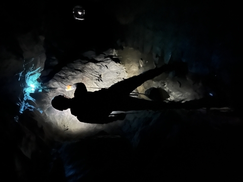 The headlamp of a researcher shines on the wall of a mine tunnel, while the UV-wand he's holding shines a turquoise-blue light on the wall above him. His profile appears, backlit by the light of his headlamp and surrounded by the darkness of the mine.