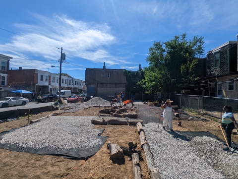 an empty lot in a city neighborhood with construction materials covering the area. Two people can be seen on the side holding tools