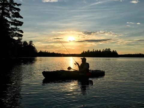 a sillhouette of a person kayak fishing while floating on a large body of water. The sun sets behind them and the sky is colored blue and yellow