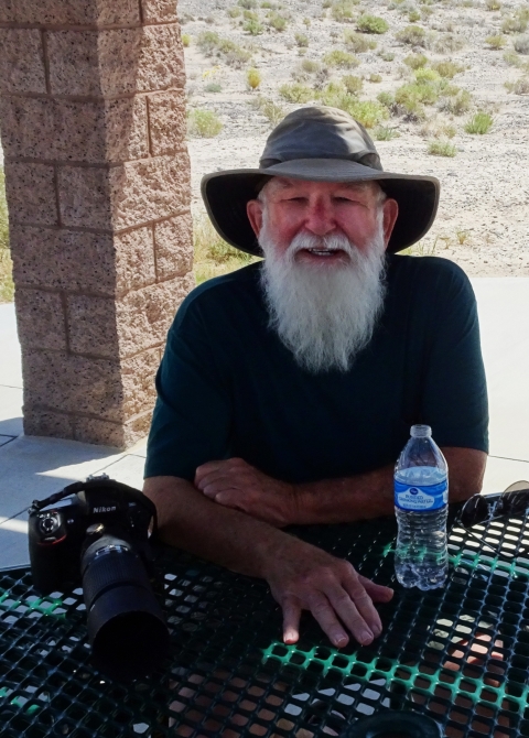 An older man with long white beard wearing a hat and dark blue shirt posing with a camera. 