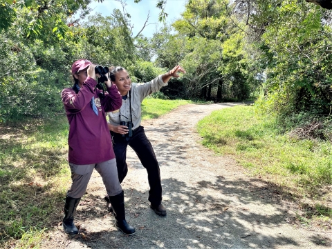Two women on a trail through woods. One of them is looking through binoculars. The other woman, in a U.S. Fish and Wildlife Service uniform, is pointing out something to the first woman.