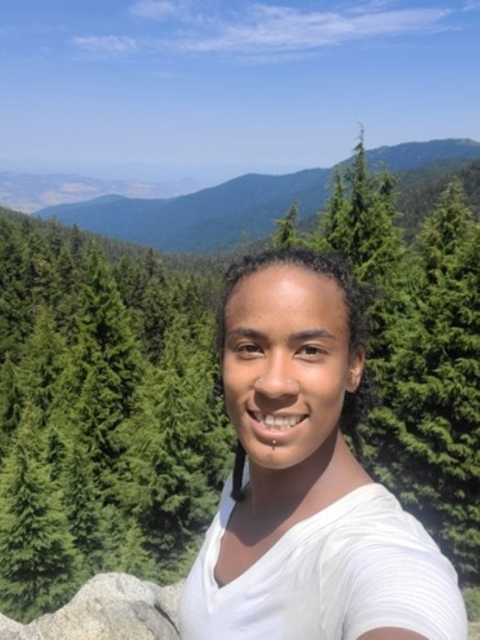 A woman smiles with a background of a forest, blue skies, and hills. 