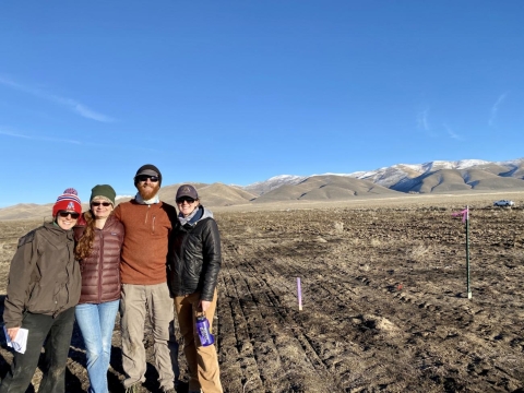 Four people standing together in front of a dirt covered landscape in winter.