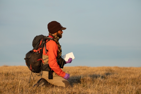 A man wearing a backpack, hiking gear, latex gloves, and a USFWS winter cap kneels in a field. In his hands he holds a notebook and a pen. 