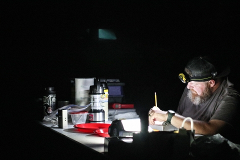 A bearded man sits in a dark environment. He has a cave lamp on his head. In front of him is a table filled with monitoring equipment. He writes in a notepad. 