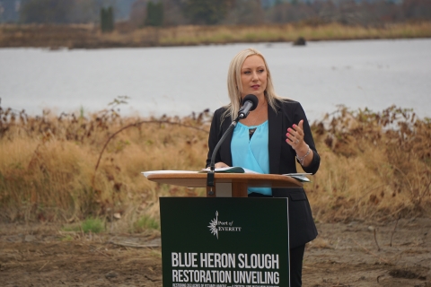 Woman with blonde hair and a blue blouse stands at a podium that says "Blue Heron Slough Restoration Unveiling"