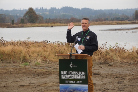 A man in a US Fish & Wildlife Service shirt stands behind a poduum that says "Blue Heron Slough Restoration Unveiling" in front of a body of water