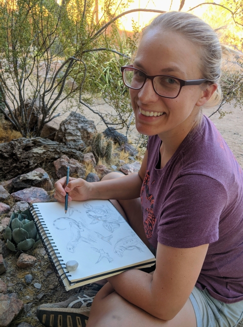 Photo of a woman in a purple shirt holding a sketchbook while sitting in a desert environment