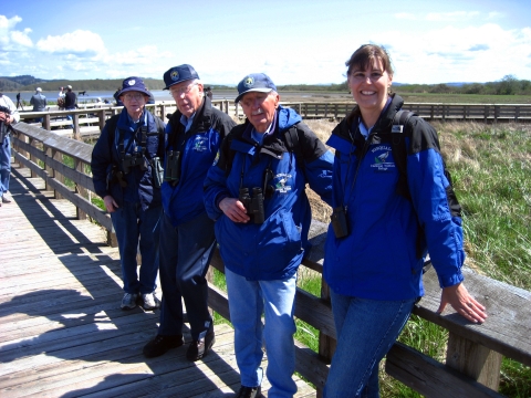 4 volunteers in blue uniforms stand together on a boardwalk trail on a sunny but chilly day, smiling into the camera.