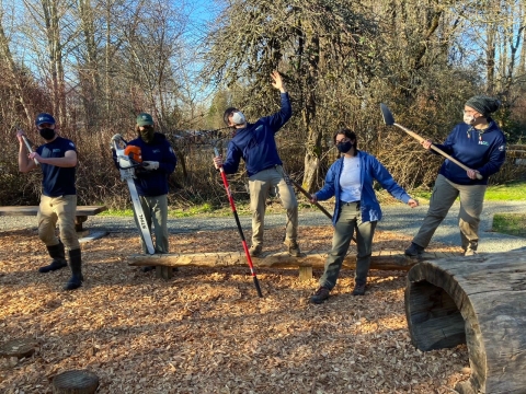 5 youth in masks and blue outerwear hold tools and pose in a play space with leafless trees in the background.