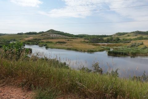 A waterway runs through a grassland that features rolling hills, shrubs, flowers, and grasses
