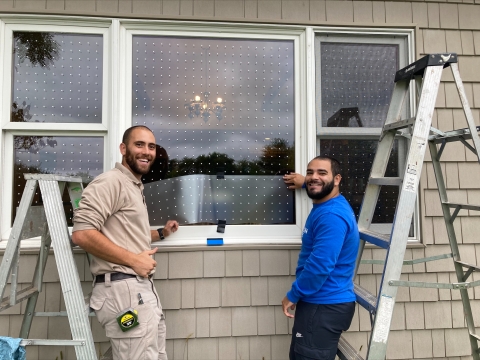 2 men hold up a transparent patterned film up to a window next to ladders