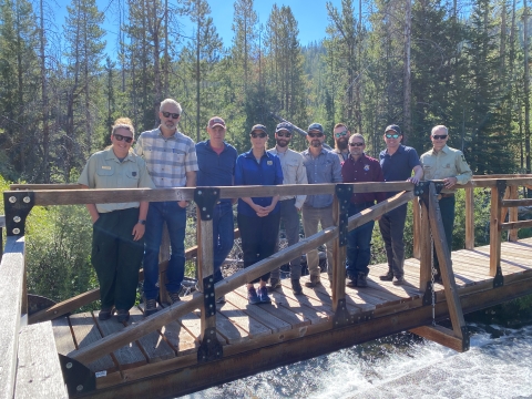 group of 10 people standing on a bridge above a fast flowing creek in the woods