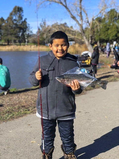 A child stands next to a body of water, holding a fish in a bag and a fishing rod