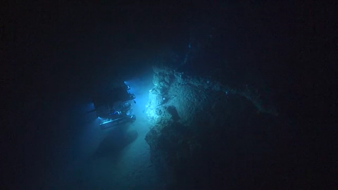 An underwater diving ship shines light on a rocky surface deep down in the ocean. The ship is surrounded by darkness