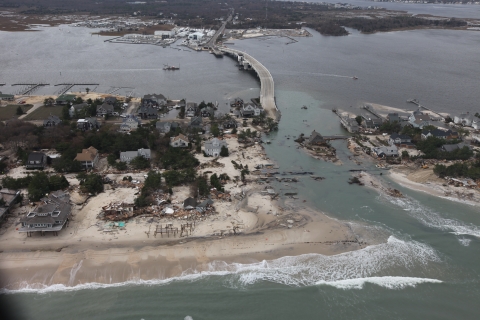 image shows an island neighborhood and houses connected to mainland by a bridge, which has been washed out by water. Debris litters the shoreline