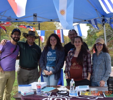 A group of people stand smiling under an outdoor tent canopy. Some wear shirts that say "Latino Outdoors" on the front. 