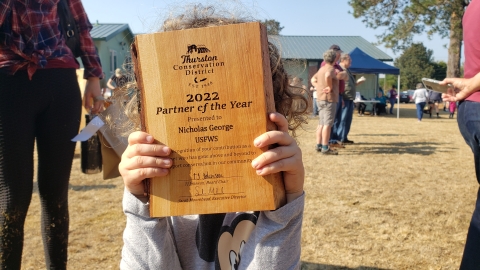 A child holds a wooden plaque in front of their face from Thurston Conservation District that says "2022 Partner of the Year, presented to Nick George, USFWS"