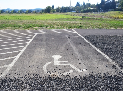 Accessible parking space with symbol for access printed in white on paved ground. Gravel parking lot surrounds it. Green field, trees, houses and mountains in background.