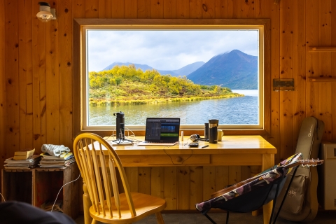 looking out the window at a lake and mountains from inside a cabin with laptop, guitar, and various items inside