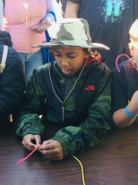 A child wearing a brimmed hat holds two colored pieces of string, tying them in a knot.