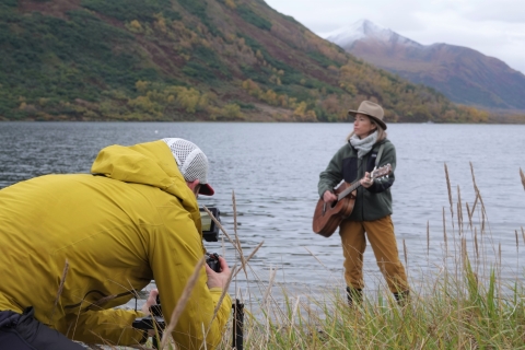 a photographer crouches to capture musician KT tunstall playing guitar in front of Karluk Lake