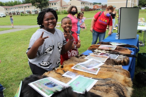 young kids smile and hold up "peace signs" next to a table with animal pelts and educational materials. A woman holding a baby and other people stand in the background