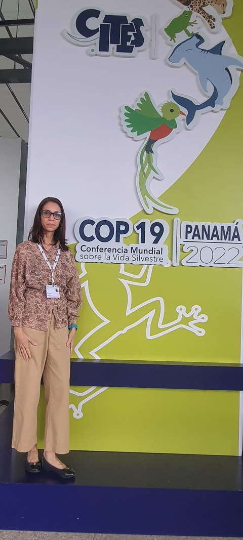 Wide shot of a female posing in front of a tall banner for CITES COP 19 in Panama, showing a hammerhead shark, jaguar, a quetzal, a pink flower, and a frog.