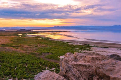 Sunset from an elevated rock outcropping, looking over a cattail marsh and large body of water. With mountains in the background. 