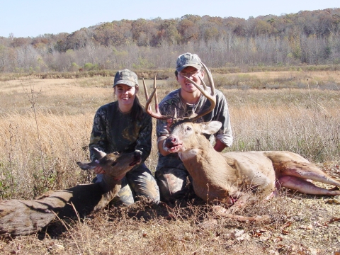 Adult man and teen girl showing their deer kills while sitting on the ground in an open field.