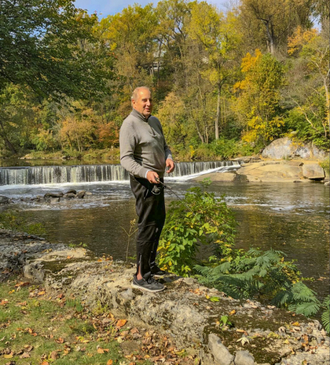 A man stands ready to cast a fishing line into a river beside him, with a short dam in the background..