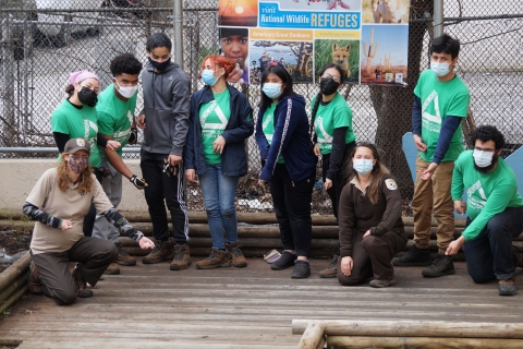 A group of people wearing face masks stand outside on a wooden platform in front of a sign that reads "Visit National Wildlife Refuges". Most of them are wearing green matching shirts with a triangular logo on the front