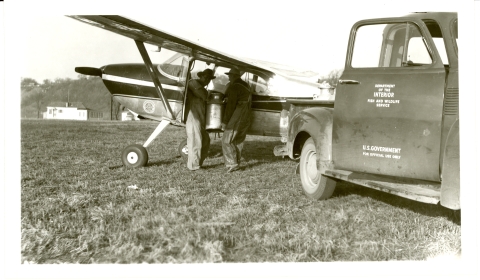 Black and white photo of milk cans with fish being loaded onto a small airplane