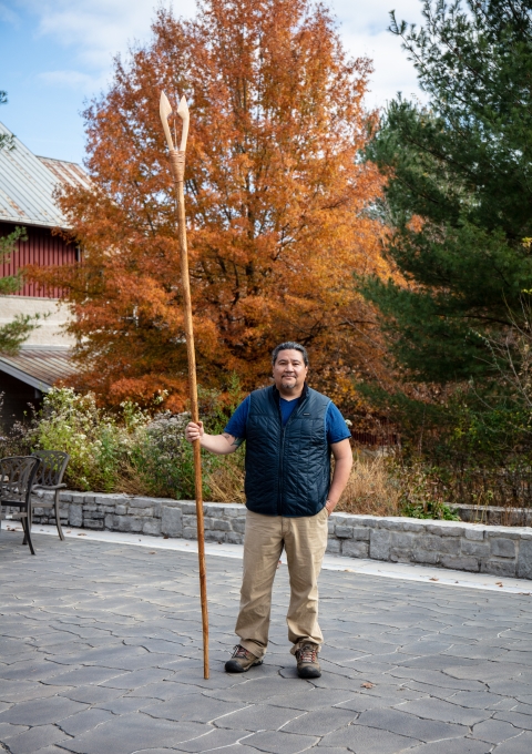 a man standing patio holding wooden spear with colorful trees in the background