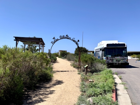 A trail that leads to the native pollinator garden as written on an overarching metal arch above the trail. Next to this is a shuttle bus.